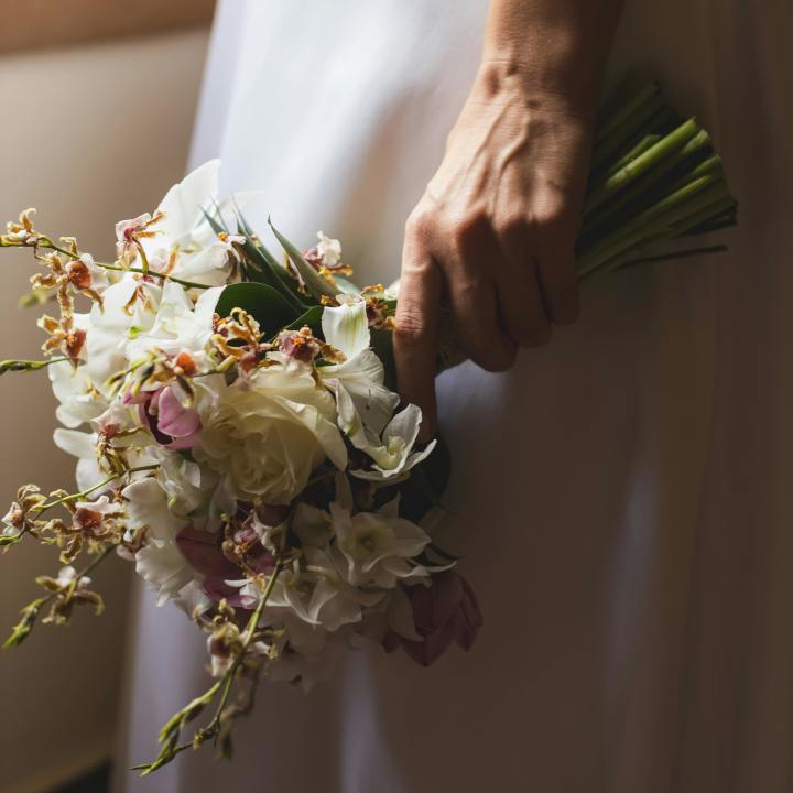 Photo of a woman in a wedding dress holding a bouquet of flowers and looking out through a window - Services - Weddings